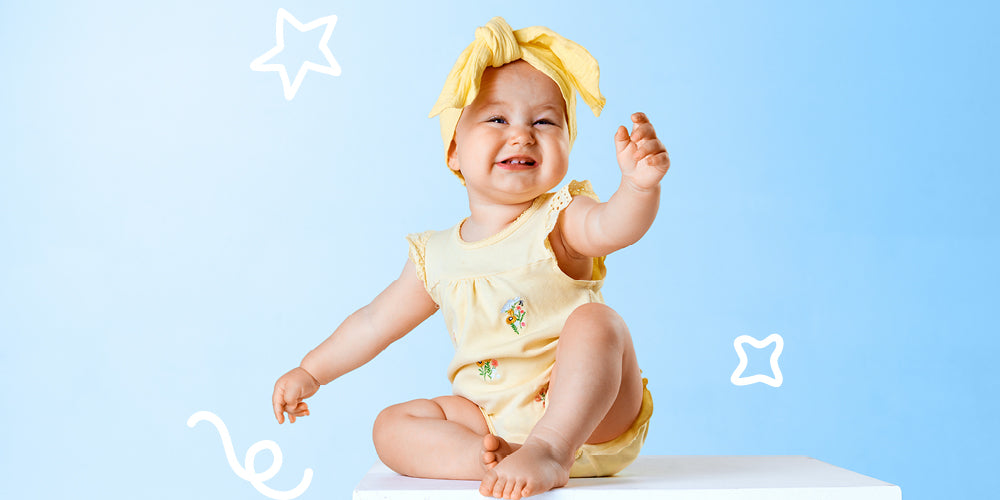 Baby in a yellow outfit and headband sitting on a white surface with a blue background