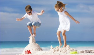Children's Orchard models on the beach, boy dressed in white polo and long jean shorts, girl dressed in white dress, both wearing sunglasses and jumping in the sand