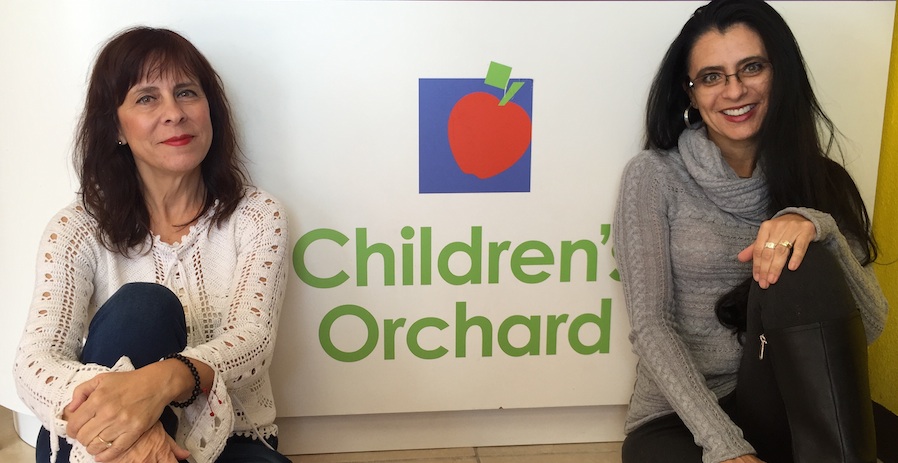 Children's Orchard franchisees Claudia and Donahi sitting on floor next to Children's Orchard logo painted on wall